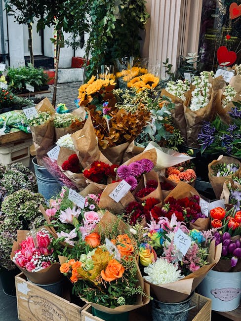 A vibrant display of fresh flowers arranged in brown paper wrapping and placed in metal buckets on a flower shop counter. The bouquets feature a variety of flowers, including bright pink tulips, orang