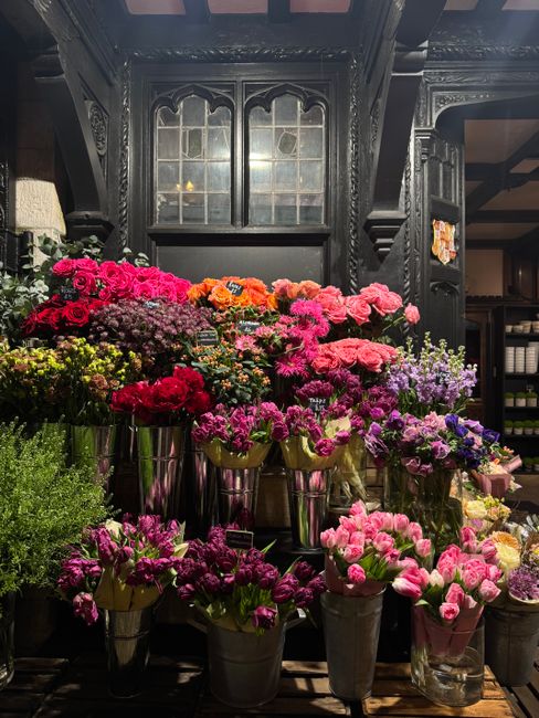 A vibrant display of fresh flowers arranged in tall, metallic vases, featuring a variety of colours including deep pinks, bright oranges, soft purples, and rich reds. The flowers include roses, carnat