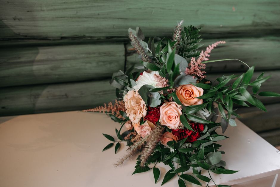 A floral bouquet featuring soft peach roses, deep red carnations, and white carnations, arranged with lush green foliage including elongated leaves, eucalyptus, and feathery astilbe. The flowers are s