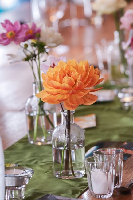 A close-up of a floral arrangement featuring a large, vibrant orange dahlia with multiple layered petals in a clear glass bottle vase, placed on a green fabric table runner. Surrounding the main flowe