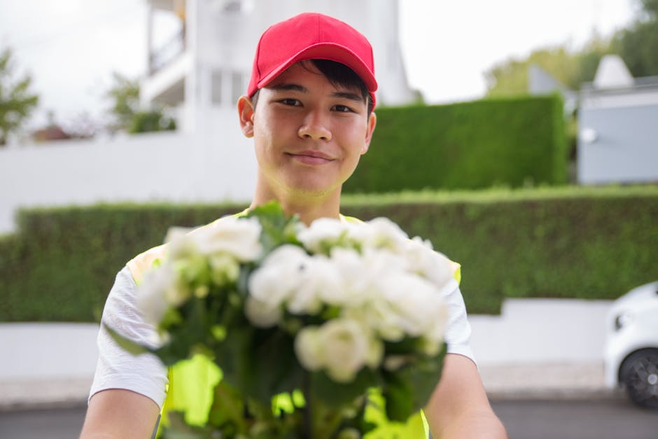 A delivery person wearing a red cap and high-visibility vest holding a bouquet of fresh white roses and greenery, extending it towards the camera with a friendly expression. The floral arrangement is