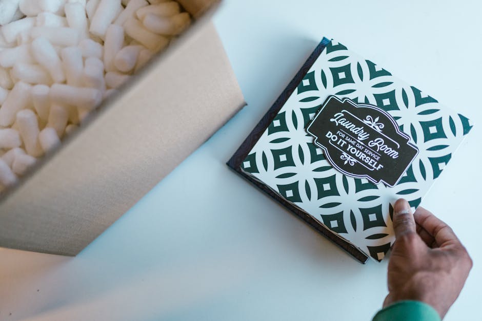 A close-up of a person's hand holding a gift box with a bold geometric black and white pattern and a label that reads 'Laundry Room for same day service do it yourself,' placed on a white surface. In